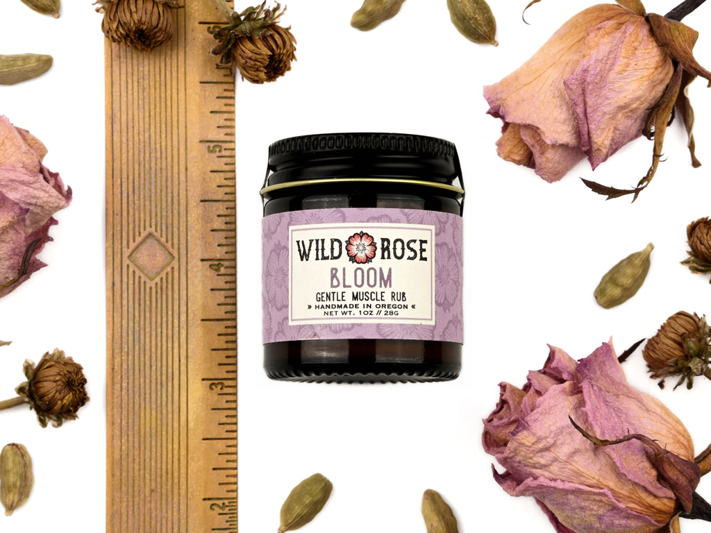 Bloom Gentle Muscle Rub in a 1oz amber glass jar with metal lid. Shown near a ruler at about 2" tall. Dried roses and cardamom pods surround.