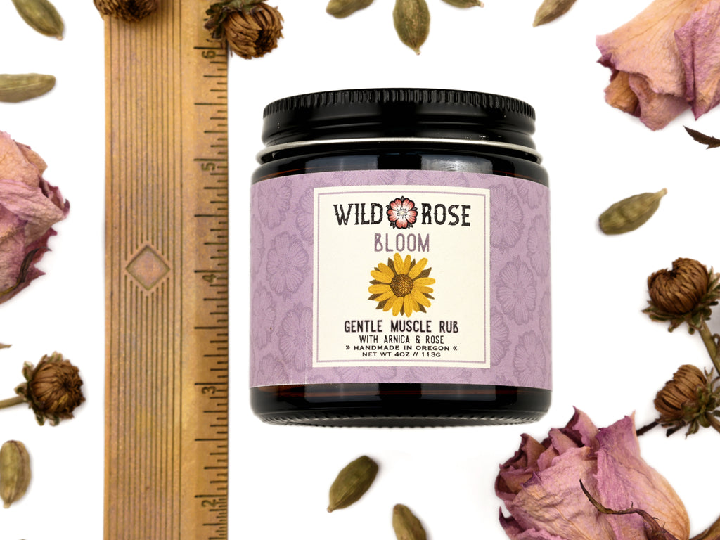 Bloom Gentle Muscle Rub in a 4oz amber glass jar with metal lid. Shown near a ruler at about 3" tall. Dried roses and cardamom pods surround.