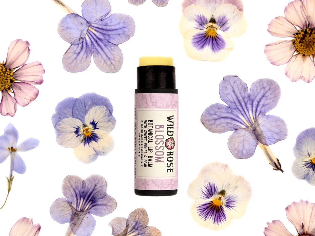 Blossom Lip Balm in a biodegradable paper tube. The cap is removed revealing a creamy light green balm. Pressed flowers surround.