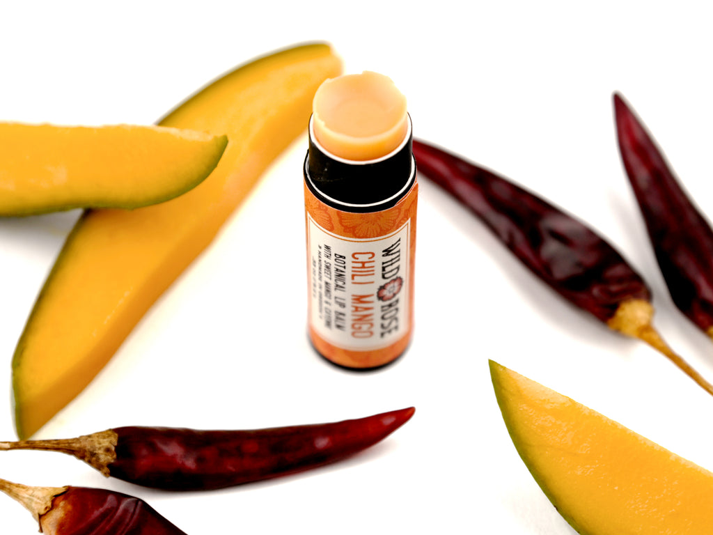 Chili Mango Natural Lip Balm in a biodegradable tube. Cap is off revealing a soft orange lip balm. Mango slices and dried chiles surround.