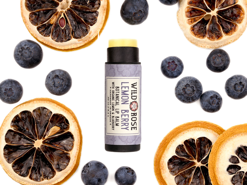 Lemon Berry Lip Balm in a biodegradable paper tube. The cap is off revealing a creamy light yellow lip balm. Dried lemon slices and blueberries surround.