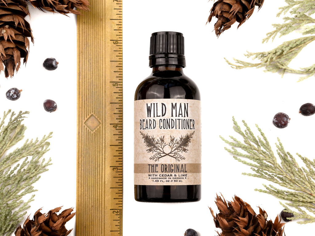 Wild Man Beard Conditioner in The Original scent shown in a 50ml amber glass bottle. Shown next to ruler measuring about 4" tall. Cedar, fir cones and juniper berries surround.