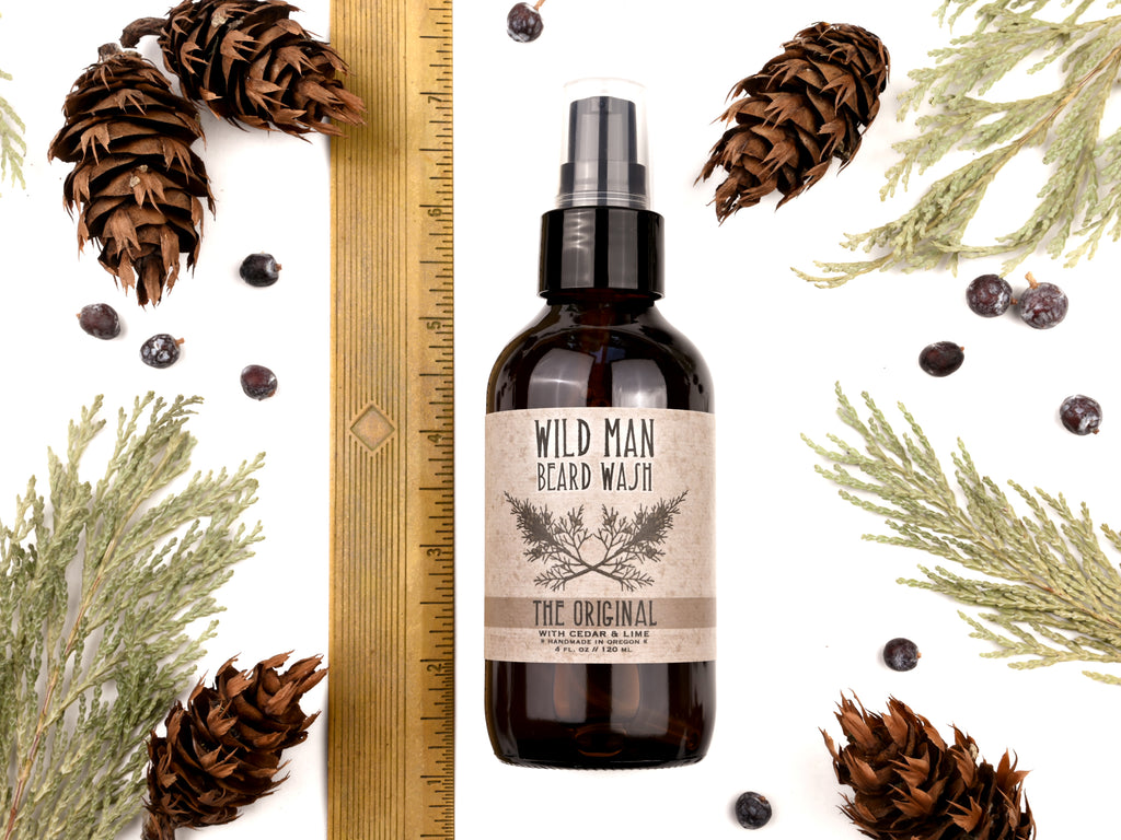 Wild Man Beard Wash in The Original scent shown in a 4oz amber glass bottle. Shown next to a ruler at about 6" tall. Cedar, fir cones and juniper berries surround.