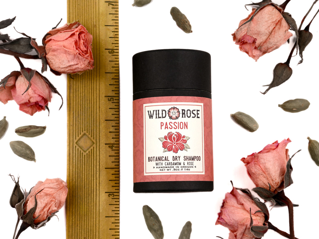 Passion Dry Shampoo in biodegradable paper shaker tube shown next to ruler at about 3" tall. Dried rose bud and cardamom pods surrounding.