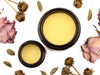 Bloom Gentle Muscle Rub in amber glass jars with lids removed revealing a creamy yellow balm. Dried flowers and cardamom pods surround.