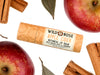 Wild Rose Apple Cider Botanical Lip Balm in a tan patterned tube, surrounded by fresh red apples and cinnamon sticks on a white background.
