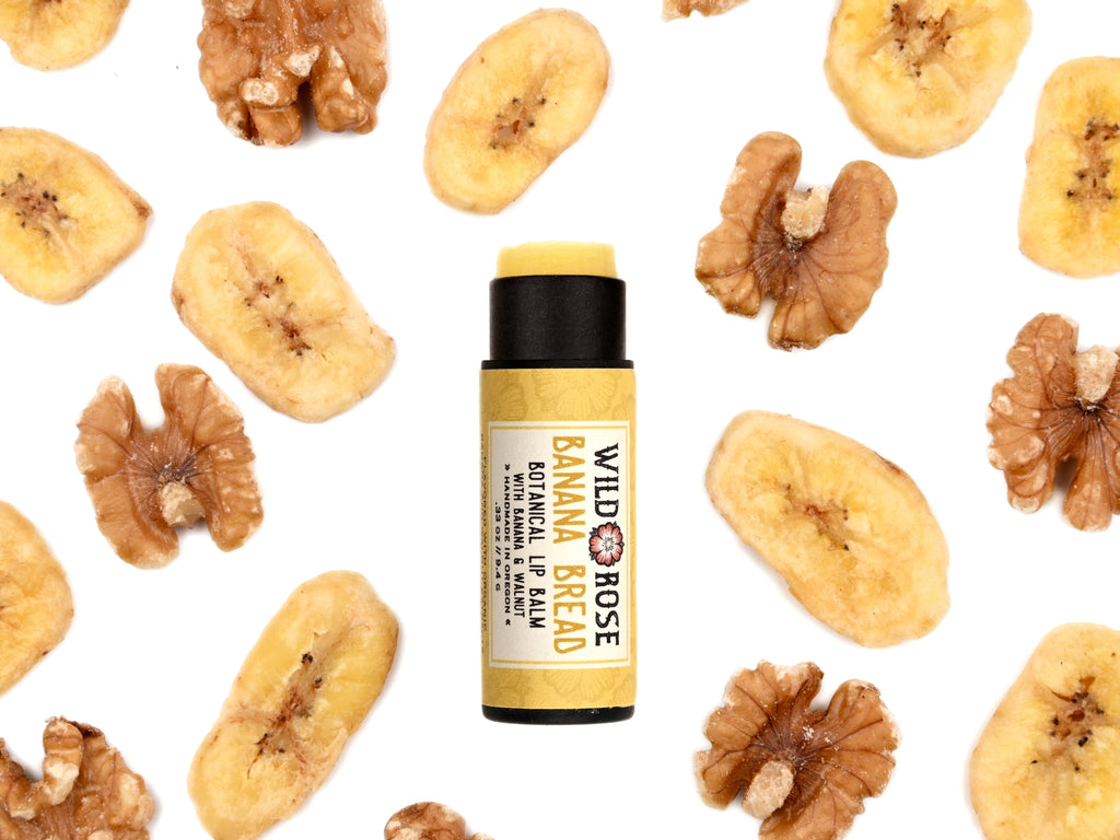 Banana Bread Natural Lip Balm in biodegradable paper tube. Cap is off revealing a creamy yellow lip balm. Dried bananas and walnuts surround.