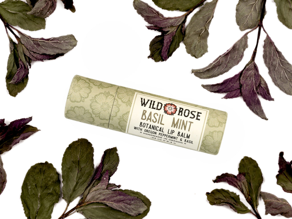 Wild Rose Basil Mint Botanical Lip Balm in a cylindrical paper tube with a floral pattern, surrounded by dried green and purple mint leaves on a white background.