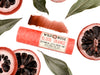 Wild Rose Blood Orange Botanical Lip & Cheek Tint lip balm tube, surrounded by sliced blood oranges and green leaves on a white background, with a product swatch above.