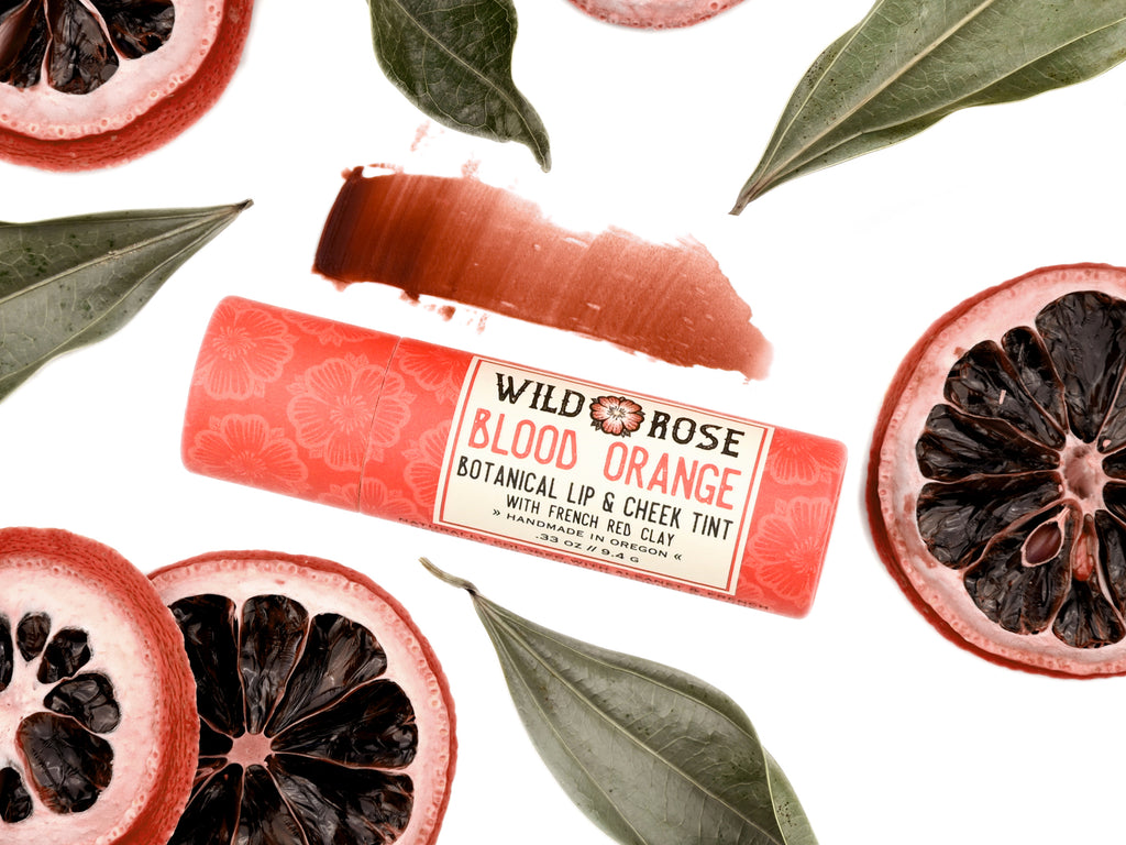 Wild Rose Blood Orange Botanical Lip & Cheek Tint lip balm tube, surrounded by sliced blood oranges and green leaves on a white background, with a product swatch above.