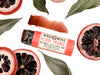 Wild Rose Blood Orange Botanical Lip & Cheek Tint lip balm tube, surrounded by sliced blood oranges and green leaves on a white background, with a product swatch above.