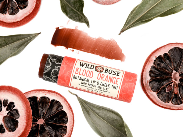 Wild Rose Blood Orange Botanical Lip & Cheek Tint lip balm tube, surrounded by sliced blood oranges and green leaves on a white background, with a product swatch above.