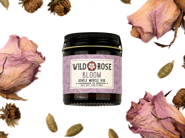 Bloom Gentle Muscle Rub in a 1oz amber glass jar with metal lid. Dried roses and cardamom pods surround.