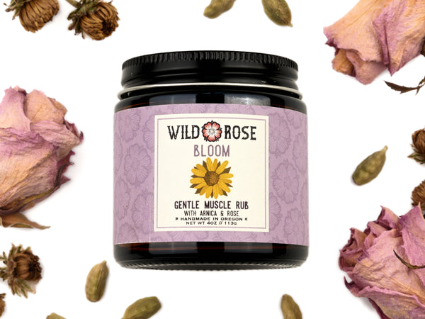 Bloom Gentle Muscle Rub in a 4oz amber glass jar with metal lid. Dried roses and cardamom pods surround.