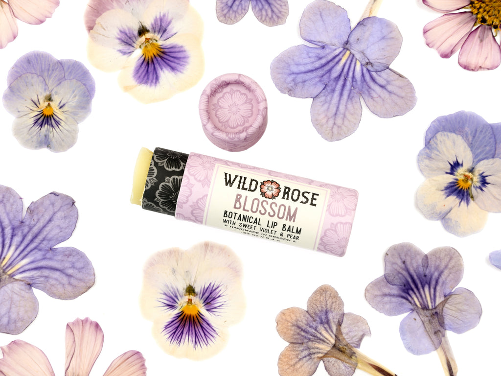 Wild Rose Blossom botanical lip balm tube placed on a white background, surrounded by  purple and lavender pansies. The lid is removed revealing a creamy, light green balm.