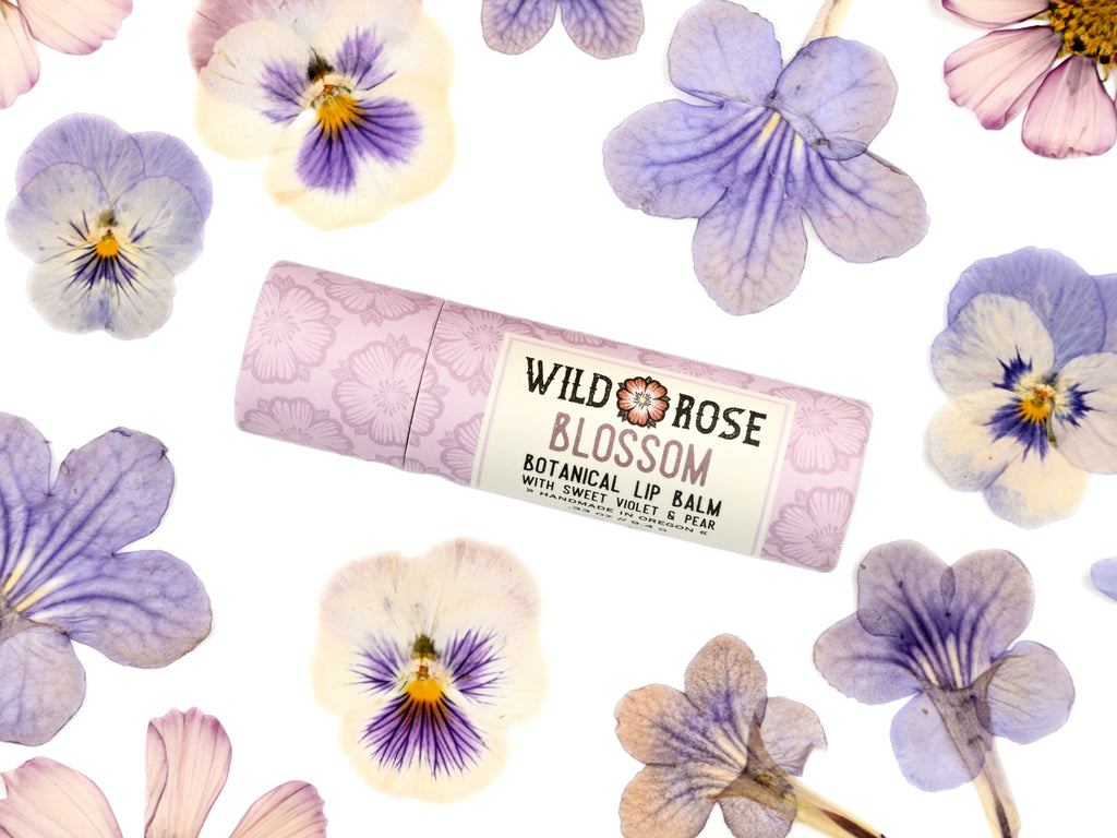 Wild Rose Blossom Botanical Lip Balm stick in a light pink patterned tube with a white label, surrounded by real purple and white pansies on a white background.