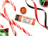 Candy Cane Natural Lip Balm in a biodegradable paper tube. Candy canes, ornaments and ribbon surround. The cap is removed, revealing a creamy white balm. 