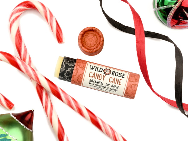 Candy Cane Natural Lip Balm in a biodegradable paper tube. Candy canes, ornaments and ribbon surround. The cap is removed, revealing a creamy white balm. 