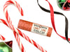 Wild Rose Candy Cane botanical lip balm tube on a white background, styled with candy canes, red and black ribbons, and green and red ornaments.