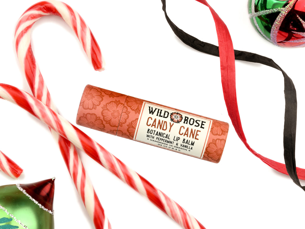 Wild Rose Candy Cane botanical lip balm tube on a white background, styled with candy canes, red and black ribbons, and green and red ornaments.