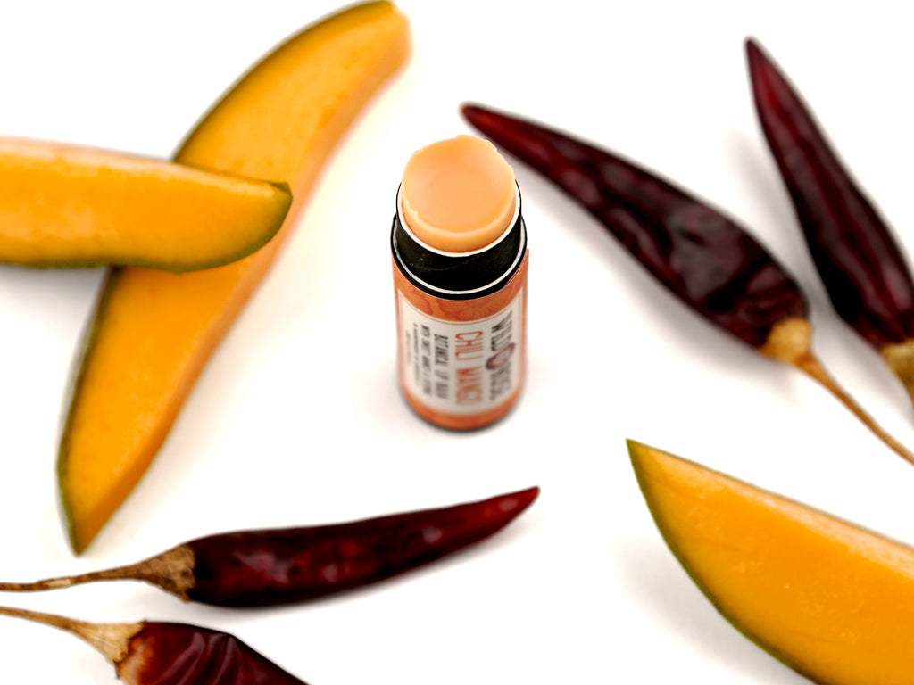 Chili Mango Natural Lip Balm in a biodegradable tube. Cap is off revealing a soft orange lip balm. Mango slices and dried chiles surround.