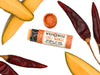 Chili Mango Natural Lip Balm in a biodegradable tube. Cap is off revealing a soft orange lip balm. Mango slices and dried chilies surround.