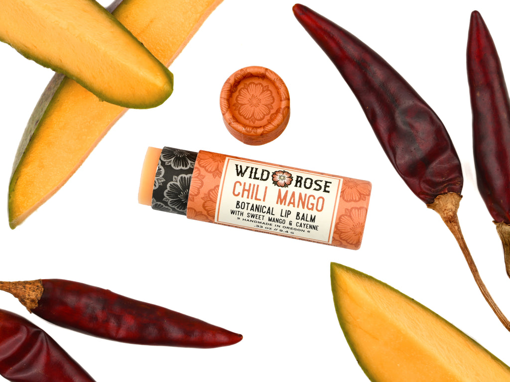 Chili Mango Natural Lip Balm in a biodegradable tube. Cap is off revealing a soft orange lip balm. Mango slices and dried chilies surround.