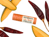Wild Rose Chili Mango Botanical Lip Balm tube centered on a white background, surrounded by sliced mango and dried red chili peppers.