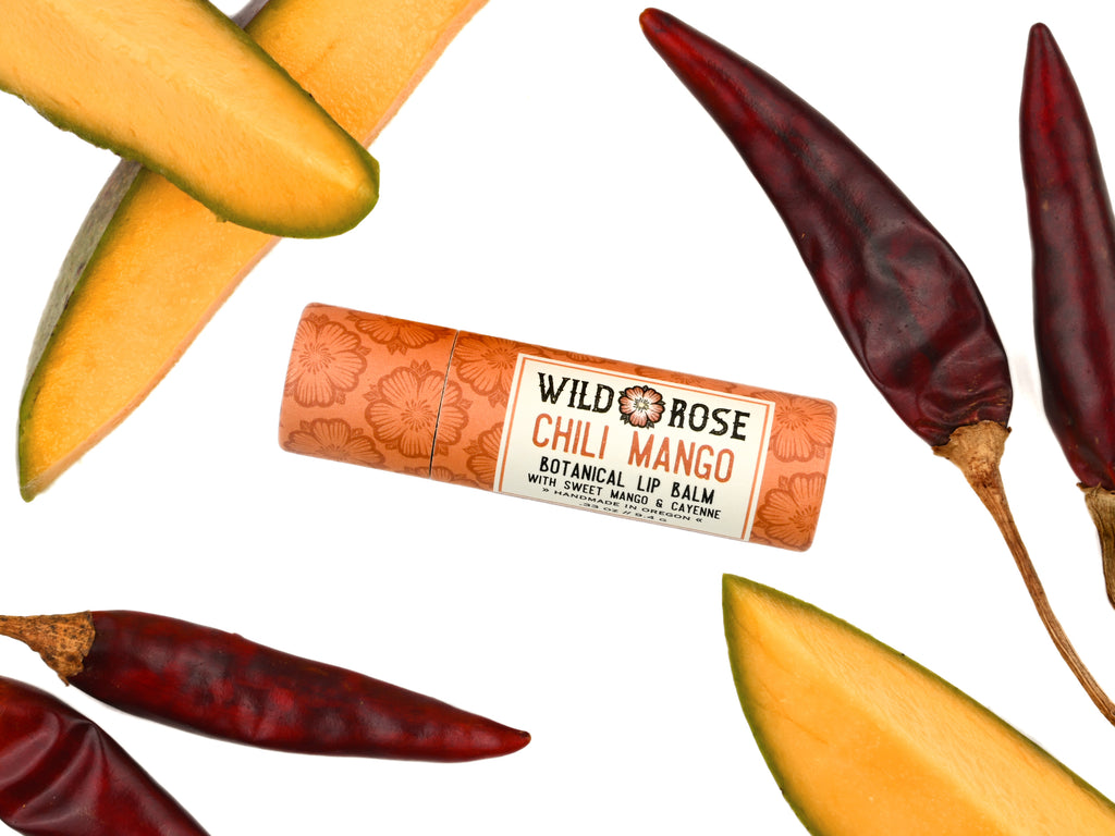 Wild Rose Chili Mango Botanical Lip Balm tube centered on a white background, surrounded by sliced mango and dried red chili peppers.