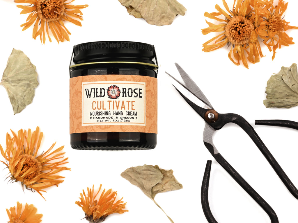 Cultivate Nourishing Hand Cream in a 1oz amber glass jar with metal lid. Shown next to tiny pruning scissors.  Dried calendula and gotu kola surround.