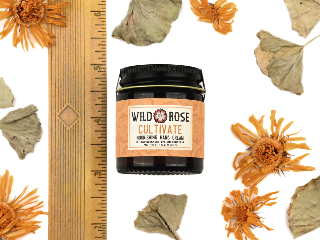 Cultivate Nourishing Hand Cream in a 1oz amber glass jar with metal lid. Shown next to ruler at about 2" tall. Dried calendula and gotu kola surround.