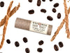 Wild Rose Dark Roast Botanical Lip Balm in a biodegradable tube, surrounded by coffee beans and vetiver on a white background. 