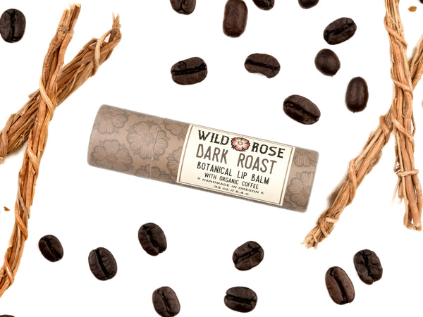 Wild Rose Dark Roast Botanical Lip Balm in a biodegradable tube, surrounded by coffee beans and vetiver on a white background. 