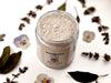 Flora Gentle Bath Salts in a glass jar. The lid is off revealing a soft, powdery bath soak within. Dried pansies and clary sage surround.