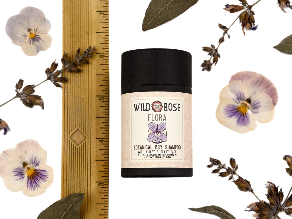 Flora Dry Shampoo in biodegradable paper shaker tube shown next to ruler at about 3" tall. Dried clary sage, pressed viola flowers and sage leaves surrounding.