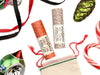 Wild Rose Holiday Lip Balm Stocking Stuffer Set with Candy Cane and Hot Cocoa Lip Balms on a white background. Ornaments and ribbon surround. Shown with a cotton gift bag with red and green stitching.