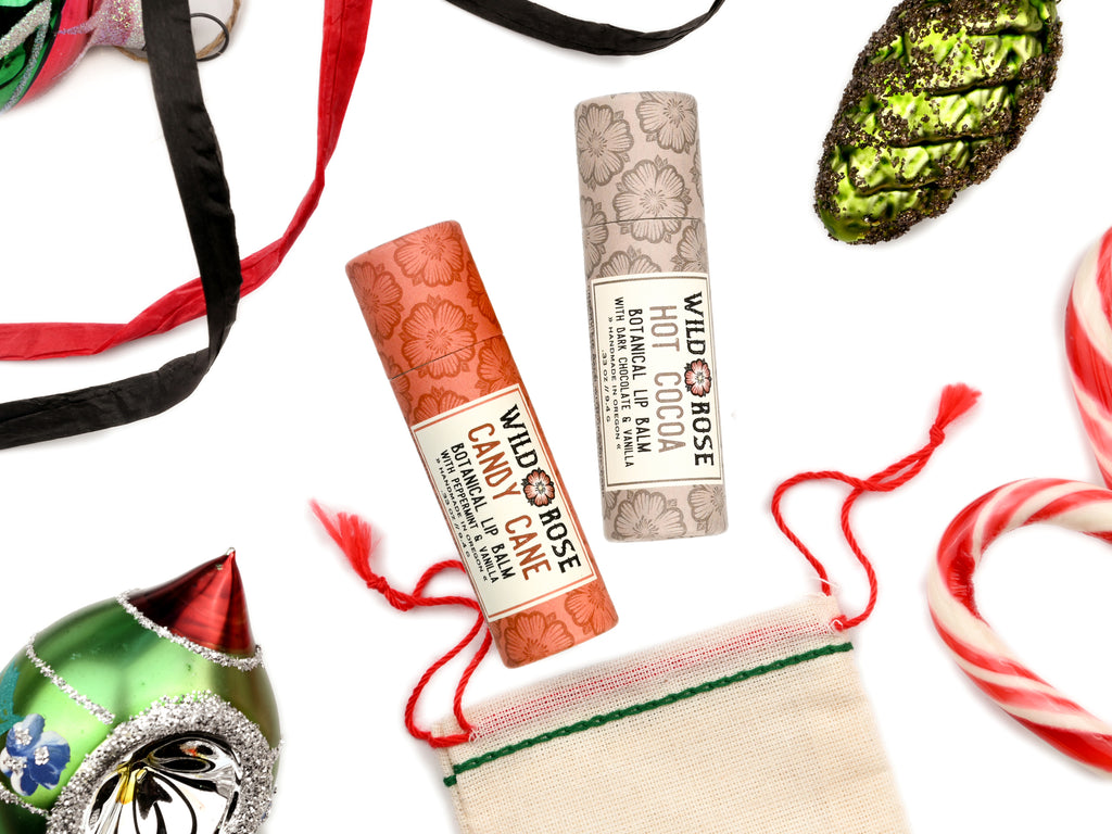 Wild Rose Holiday Lip Balm Stocking Stuffer Set with Candy Cane and Hot Cocoa Lip Balms on a white background. Ornaments and ribbon surround. Shown with a cotton gift bag with red and green stitching.