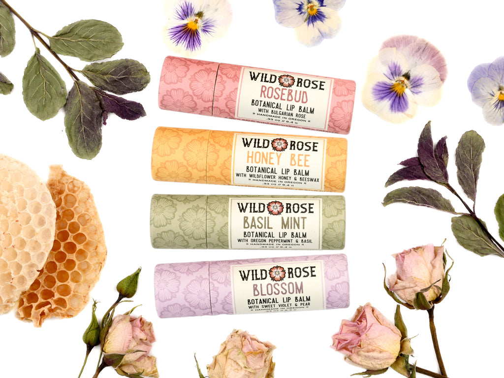 Wild Rose Garden Lip Balm Set with Rosebud, Honey Bee, Basil Mint and Blossom Lip Balms in biodegradable tubes. Shown with honeycomb, dried mint, dried rosebuds and pressed pansies on a white background. 