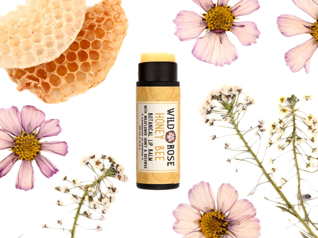 Honey Bee Natural Lip Balm in a biodegradable paper tube shown with cap off revealing a creamy yellow balm. Honey comb and pressed flowers surround.