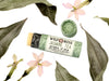 Jasmine Tea Natural Lip Balm in a biodegradable paper tube. The cap is off, revealing a creamy off-white balm. Dried leaves and pressed flowers surround.
