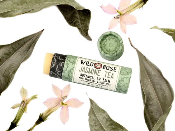 Jasmine Tea Natural Lip Balm in a biodegradable paper tube. The cap is off, revealing a creamy off-white balm. Dried leaves and pressed flowers surround.