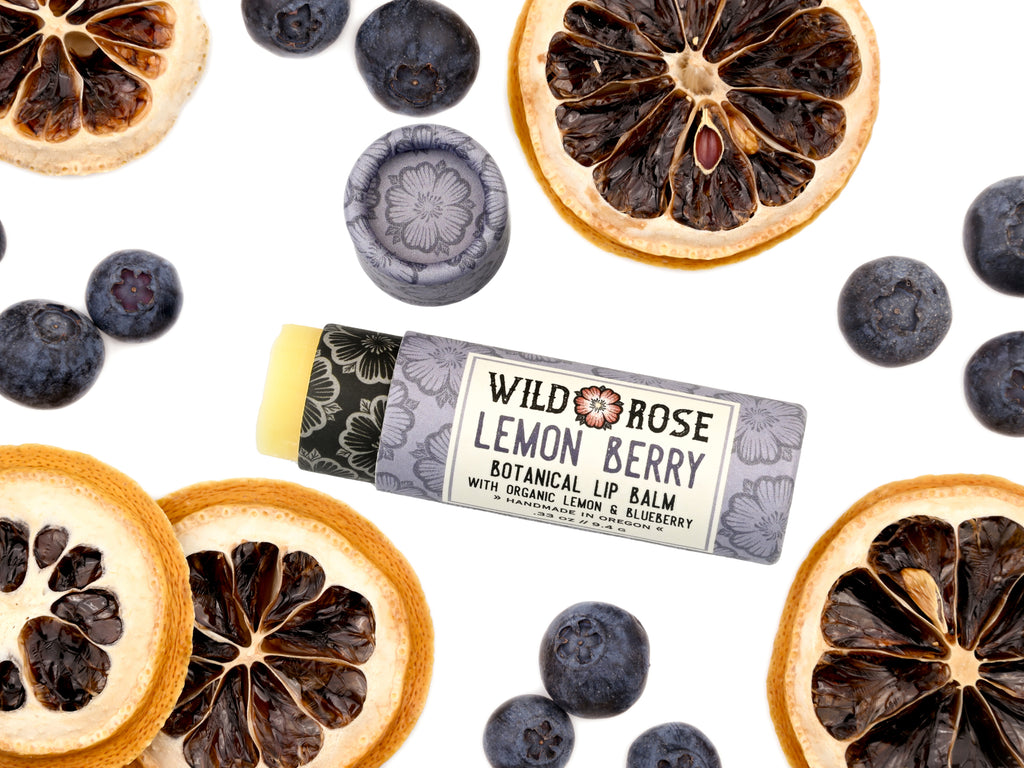 Wild Rose Lemon Berry Botanical Lip Balm in a biodegradable tube on a white background with dried lemons and blueberries surrounding. The cap is removed revealing a creamy yellow balm.