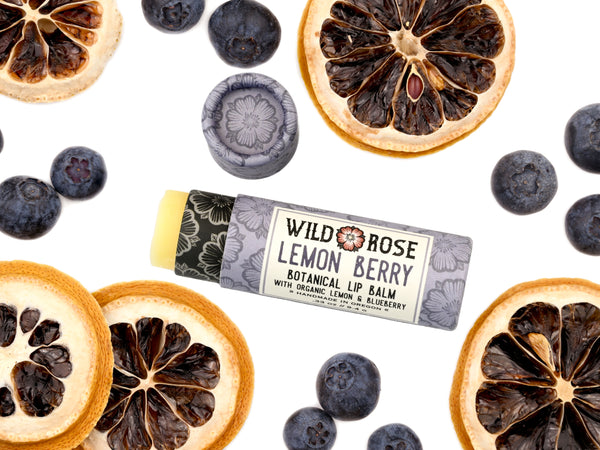 Wild Rose Lemon Berry Botanical Lip Balm in a biodegradable tube on a white background with dried lemons and blueberries surrounding. The cap is removed revealing a creamy yellow balm.
