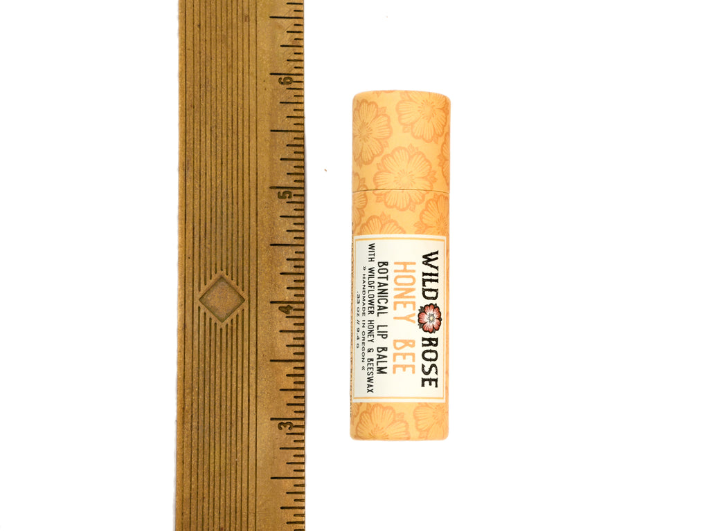 A tube of Wild Rose Honey Bee Botanical Lip Balm in a biodegradable paper tube stands next to a ruler on a plain white background, showing its size.