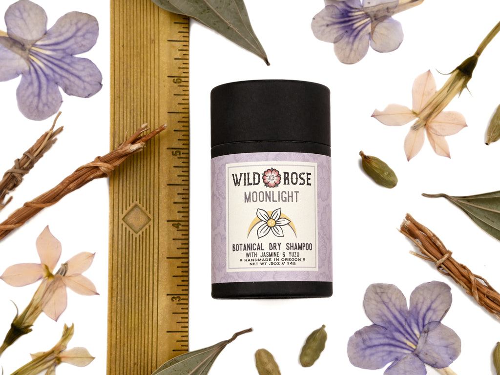 Moonlight Dry Shampoo in biodegradable paper shaker tube shown with ruler at about 3" tall. Jasmine, vetiver, cardamom and purple flowers surrounding.