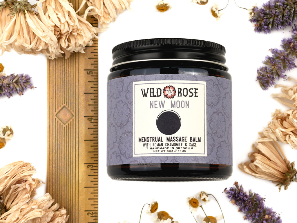 New Moon Menstrual Massage Balm in a 4oz amber glass jar with metal lid. Shown near a ruler at about 3" tall.  Dried flowers surround.