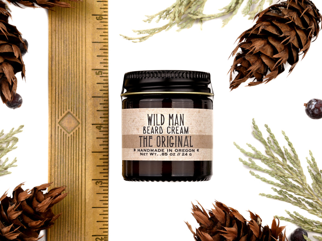 Wild Man Beard Cream in The Original scent shown in a 1oz amber glass jar. Shown next to a ruler at about 2" tall. Cedar, fir cones and juniper berries surround.