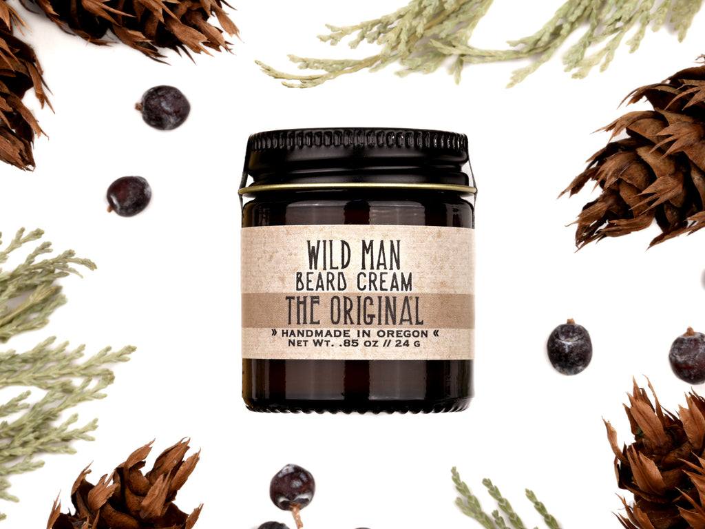 Wild Man Beard Cream in The Original scent shown in a 1oz amber glass jar. Cedar, fir cones and juniper berries surround.