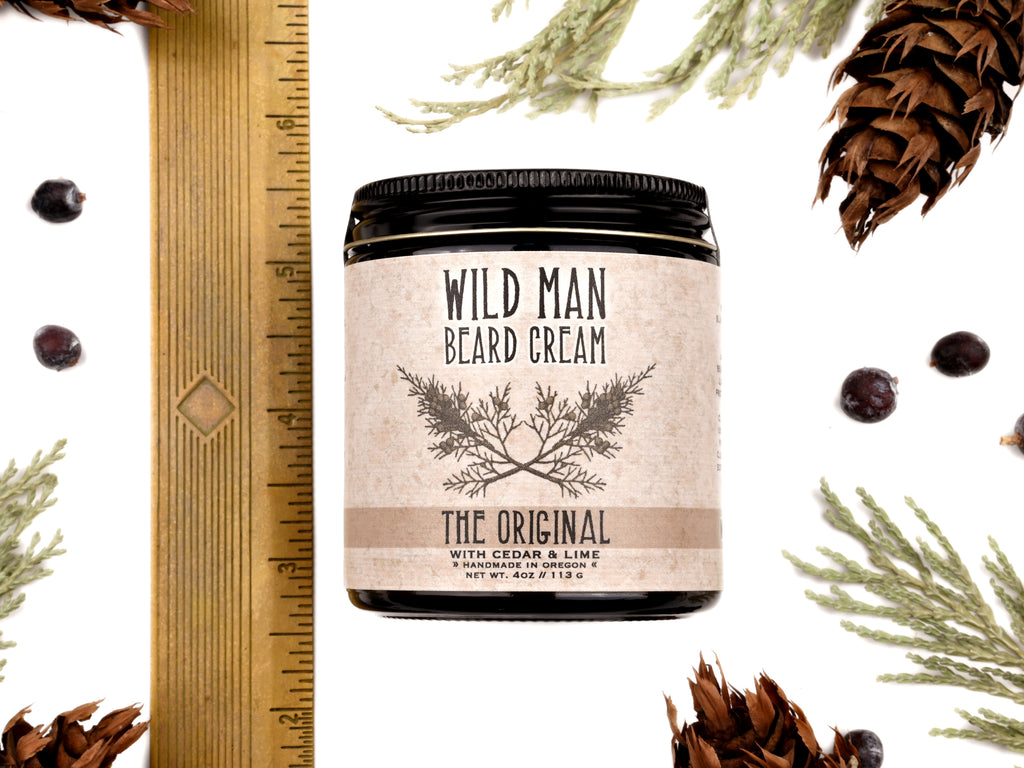 Wild Man Beard Cream in The Original scent shown in a 4oz amber glass jar. Shown next to a ruler at about 3" tall. Cedar, fir cones and juniper berries surround.
