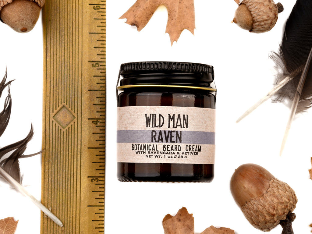 Wild Man Beard Cream - Raven scent in 1oz amber glass jar. Shown next to ruler at about 2" tall. Black feathers, acorns and dried oak leaves surround. 